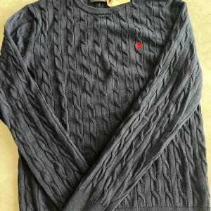 Kabelstickad tröja från Morris  - Kabelstickad marinblå tröja från Morris i merinoull. Klassisk rund halsringning, långärmad och med röd broderad logga på bröstet. Inga defekter. Storlek M. Pris 500kr