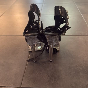 Svarta platåklackar med 15cm klack och snörning - Säljer ett par svarta platåklackar från Shuzumiao i storlek 36. Skorna har 15cm höga genomskinliga klackar och transparent platåsula. Ovandelen är i lackmaterial med snörning och metallspännen på sidorna. Öppen tå och cool statement-look.