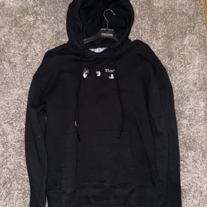 Svart Off-White hoodie med tryck - Svart hoodie från Off-White med vit logga och handmotiv framtill. Baksidan har ett stort, färgstarkt tryck i rött, orange och grått som bildar ett unikt mönster. Tröjan har huva med snörning och en klassisk känguruficka. Perfekt för dig som gillar streetwear och statement-plagg.