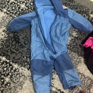 Blå overall med fleecefoder, stl 86 - Blå overall med huva och dragkedja framtill. Overallen har mönstrade partier i blått och är fodrad med mjuk fleece på insidan. Perfekt för kyliga dagar och enkel att ta på tack vare dragkedjan. Elastiska muddar vid ärmar och ben för extra komfort.