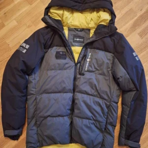 Grå och svart pufferjacka Sail Racing - Snygg pufferjacka från Sail Racing i grått och svart med gul insida. Jackan har huva, dragkedja framtill och logga på bröstet samt ärmarna. Perfekt för kalla dagar när du vill hålla stilen och värmen. Sällan använd och är i extremt bra skick