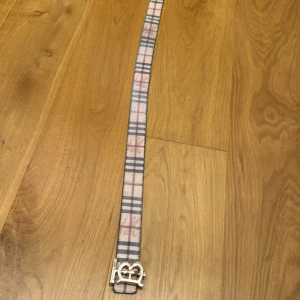 Burberry bälte med rutigt mönster - Säljer ett klassiskt Burberry bälte med ikoniskt rutigt mönster och en stor, guldig B-spänne framtill. Bältet finns i både beige och svart variant, båda med tydliga rutor och exklusiv känsla. Perfekt accessoar för att lyfta din outfit med en lyxig touch.