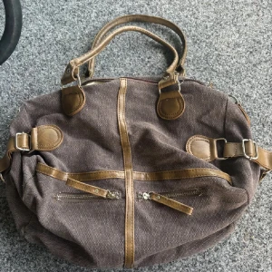 Brun handväska i canvas och skinn - Rymlig brun handväska i canvas med detaljer och handtag i brunt skinn. Väskan har två dragkedjefickor framtill, justerbara spännen på sidorna och ett ljust, glansigt innerfoder med innerficka. Perfekt för dig som vill ha en snygg och praktisk väska till vardagen. Endast swish betalning skickas spårbart 59kr