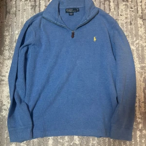 Blå half zip tröja från Polo Ralph Lauren - Säljer en blå långärmad tröja från Polo Ralph Lauren med gul broderad logga på bröstet. Tröjan har en klassisk half zip-dragkedja och är tillverkad i mjuk bomull. Perfekt för lager-på-lager och chill stil.
