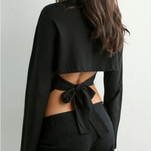 Svart långärmad topp med öppen rygg - Snygg svart långärmad topp med öppen rygg och knytning bak. Toppen har en croppad passform och är tillverkad i ett mjukt material som sitter skönt mot kroppen. Perfekt för dig som vill ha en stilren men ändå unik look.