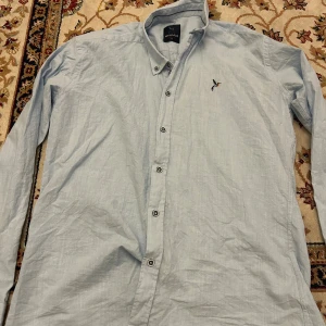 Ljusblå skjorta från Megalith - Snygg ljusblå skjorta från Megalith med klassisk krage och broderad fågel på bröstet. Skjortan har långa ärmar och knäppning framtill med svarta knappar. Perfekt för en clean och stilren look.