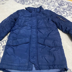 Blå pufferjacka från H&M, stl 134 - Snygg mörkblå pufferjacka från H&M med hög krage och stora fickor framtill. Jackan har dragkedja och tryckknappar, samt en extra ficka med dragkedja på ärmen. Perfekt för kalla dagar och enkel att matcha med det mesta.