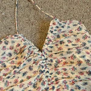 Blommig kortklänning från Zara XS - Supersöt kort klänning från Zara i blommigt mönster med volangkant nertill och smala axelband. Klänningen har rynkade detaljer framtill och är i en ljus, krämig ton med rosa, blå och gröna blommor. Perfekt för sommarens alla tillfällen!