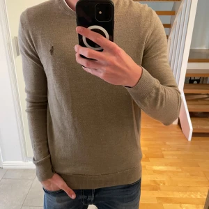 Beige stickad tröja från Polo Ralph Lauren - Säljer en stilren beige stickad tröja från Polo Ralph Lauren. Tröjan har rund halsringning, diskret logga broderad på bröstet och långa ärmar. Perfekt för lager-på-lager och passar till jeans eller chinos. (Modell 176cm, 70kg)
