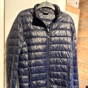 Mörkblå dunjacka från Emporio Armani - Snygg mörkblå dunjacka från Emporio Armani EA7 med quiltad design och dragkedja framtill. Jackan har hög krage, två sidofickor och EA7-logga på bröstet. Perfekt för kyliga dagar och ger en sportig vibe. Dragkedjan kan gå ut ibland men är enkel att justera.