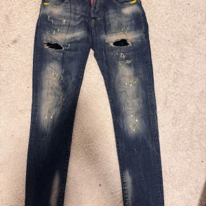Dsquared2 slim jeans med färgstänk - Snygga blå slim fit jeans från Dsquared2 med slitna detaljer, hål på knäna och coola färgstänk i gult och vitt. Klassisk femficksmodell med Dsquared2-logga vid fickan. Tillverkade i Italien av bomull.