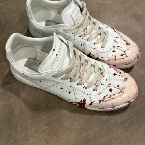Maison Margiela sneakers med röd färg - Unika vita sneakers från Maison Margiela i skinn med röda färgstänk över hela skon. Klassisk låg modell med snörning och rund tå. Snygg och edgy design som sticker ut, perfekt för dig som gillar statement pieces.
