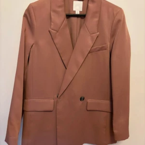 Rosa kavaj från H&M i XS - Snygg rosa kavaj från H&M i storlek XS. Dubbelknäppt med svarta knappar, klassisk krage och fickdetaljer framtill. Stilren och modern passform, perfekt för dig som gillar att sticka ut med färg. Materialet är mjukt och har en lätt glansig finish.