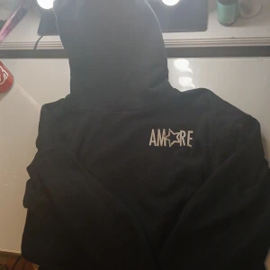 Svart hoodie från UNIQ Studio med tryck - Svart hoodie från UNIQ Studio med stort vitt AM★RE-tryck på bröstet. Klassisk modell med huva och mjukt material, perfekt för chill dagar. Snygg och enkel att matcha med jeans eller joggers.