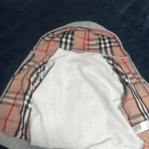 Grå hoodiejacka från Burberry M - Grå hoodiejacka från Burberry med dragkedja, klassiskt rutigt foder i beige, svart och rött samt broderad logga på bröstet. Jackan har huva med snörning och ribbade muddar. Perfekt för dig som vill ha en stilren och lyxig streetwear-look, säljer varan eftersom den var för liten för mig så för den mesta delen har den legat hemma orörd. nu tänker jag det är dags att sälja den vidare.