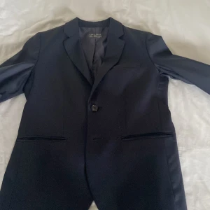 Mörkblå kavaj från Zara Boys 152 - Snygg mörkblå kavaj från Zara Boys i storlek 152. Klassisk modell med en knapp framtill, två fickor och slät finish. Perfekt för dig som vill ha en stilren och enkel look. Fodrad insida och diskreta detaljer.