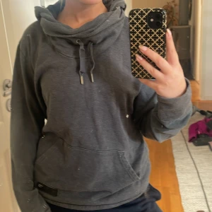 Mörkgrå hoodie med snörning - Mysig mörkgrå hoodie med stor huva och snörning i halsen. Tröjan har en känguruficka framtill och långa ärmar. Perfekt för chill dagar eller när du vill vara extra bekväm.