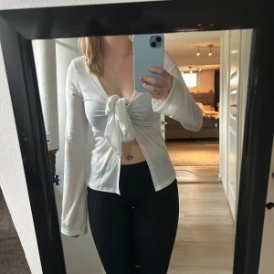 Vit långärmad knytblus  - Säljer en vit långärmad blus. Toppen har en djup v-ringning och knyts framtill med en stor rosett. Tunt och mjukt material som känns skönt mot huden. Perfekt för dig som gillar trendiga och feminina detaljer.