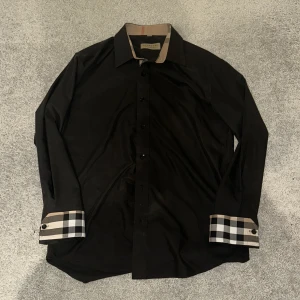 Svart Burberry skjorta med rutiga detaljer - M - passar som S. Svart skjorta från Burberry med klassiska rutiga detaljer i beige, svart och vitt på krage och manschetter. Skjortan har lång ärm, knappar framtill och en stilren, normal passform. Perfekt för dig som vill ha en clean look med en touch av lyx.