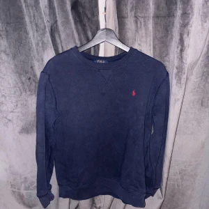 Mörkblå tröja från Polo Ralph Lauren - En stilren mörkblå tröja från Polo Ralph Lauren med klassisk rund halsringning och röd broderad logga på bröstet. Tröjan har långa ärmar och är tillverkad i mjuk bomull, perfekt för en avslappnad och snygg look.