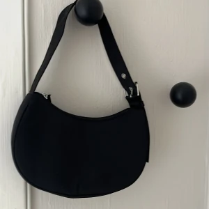 Svart axelväska med justerbart band - Snygg svart axelväska i minimalistisk design med rundad form. Väskan har ett justerbart axelband med silvriga metalldetaljer och dragkedja upptill. Perfekt storlek för det viktigaste och passar till många olika stilar.