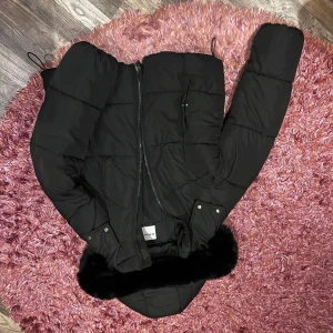 Svart pufferjacka från Bershka - Säljer en svart pufferjacka från Bershka i storlek L men känns mera som M. Jackan har en mysig huva med fejkpäls, dragkedja framtill och två fickor med tryckknappar.andvänd en halv vinter, köpt för 699kr 