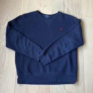 Mörkblå sweatshirt från Polo Ralph Lauren - Klassisk mörkblå sweatshirt från Polo Ralph Lauren med rund halsringning och röd broderad logga på bröstet. Tröjan har långa ärmar och ribbade muddar vid ärmslut och nederkant. Perfekt för en chill och stilren look.