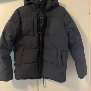 Svart pufferjacka från Five Seasons - Svart pufferjacka från Five Seasons med huva och dragkedja. Jackan har två stora fickor framtill och ett camouflagemönstrat innerfoder. Materialet är polyester och jackan är både vind- och vattentät. Perfekt för kalla dagar när du vill hålla stilen.