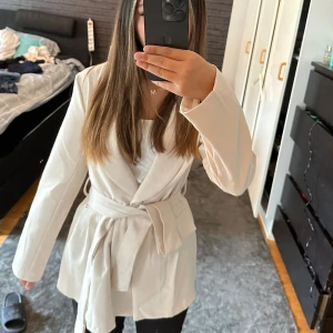 Beige kavaj med bälte - Snygg beige kavaj med omlott och knytbälte i midjan. Jackan har långa ärmar och en stilren, klassisk krage. Perfekt för dig som vill ha en clean och trendig look. Passar till många olika outfits och ger en fräsch vibe. XSS-S