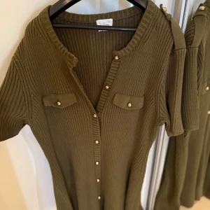  ribbad klänning med knappar - Snygg olivgrön ribbstickad klänning från HM med kort ärm, guldknappar framtill och dekorativa fickor på bröstet. Klänningen har en v-ringad hals och är i mjukt stickat material som ger en skön passform. Aldrig använd pga stor storlek