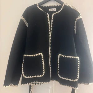 Svart cardigan jacka från VERO MODA - Svart cardigan jacka från vero moda  i storlek XS. Jackan har kontrasterande vita broderade kanter, två stora fickor framtill och ett matchande knytbälte i midjan. Rak passform och långärmad, perfekt. Materialet är lite luddigt, men det går lätt att fixa med en noppborttagare. Annars i fint skick! 💫