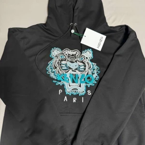 Svart Kenzo hoodie med tigerbrodyr - Svart hoodie från Kenzo med ikonisk tigerbrodyr i vitt, blått och turkost framtill. Tröjan har huva med snörning och är tillverkad i 100% bomull. Klassisk passform och långärmad modell, perfekt för en cool streetstyle-look.