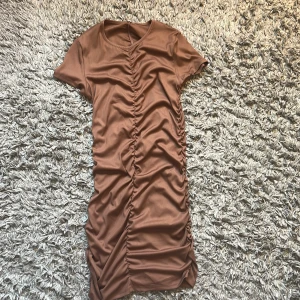 Brun kortklänning med snörning - Säljer en brun kortklänning med snygg rynkad snörning längs hela mitten, både fram och bak. Klänningen har korta ärmar och en rund halsringning. Materialet känns mjukt och stretchigt, perfekt för en trendig look.