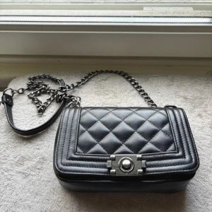 Svart quiltad axelväska i skinn - Snygg svart axelväska i äkta skinn från Borse in Pelle, tillverkad i Italien. Väskan har quiltad design, silverfärgad kedjerem och coolt lås framtill. Perfekt storlek för det viktigaste och ger en lyxig vibe till din outfit. Insidan är lite smutsig men inget man tänker på. Spännet kan vara lite segt ibland.