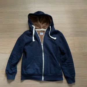 Mörkblå hoodie med fur - Mysig mörkblå hoodie med dragkedja och vit snörning. Insidan av huvan är fodrad med brunt teddyfoder för extra värme. Perfekt för kyliga dagar och har två stora fickor framtill.