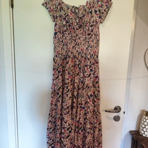 Blommig långklänning från John Baner stl 38 - Supersöt långklänning från John Baner med blommigt mönster i rosa, blått och brunt på ljus botten. Klänningen har korta ärmar, smock över bysten och knytning vid halsen. Perfekt för varma dagar och ger en bohemisk vibe.