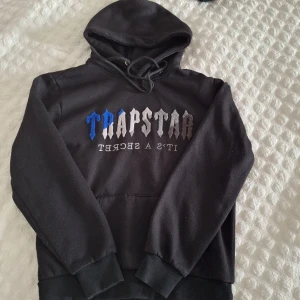 Svart Trapstar hoodie med brodyr - Svart hoodie från Trapstar med broderad logga i blått, vitt och grått på bröstet. Klassisk huva med snörning och stor magficka framtill. Mjuk insida och ribbade muddar vid ärmslut och nederkant. Perfekt för en streetwear-look.