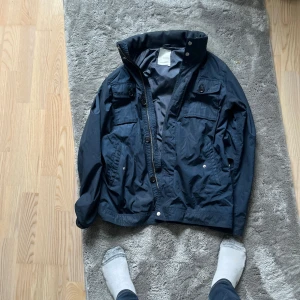 Mörkblå vindjacka från J.Lindeberg - Snygg mörkblå vindjacka från J.Lindeberg med hög krage, flera fickor framtill och både dragkedja och knappar. Jackan har en klassisk passform och är perfekt för blåsiga dagar. Materialet är lätt och smidigt, vilket gör den enkel att bära.