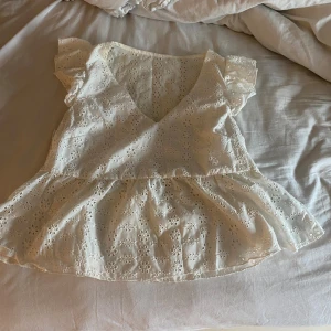 Vit blus med volang och broderade blommor - Supersöt vit blus med v-ringning, volang nertill och korta volangärmar. Tunt bomullstyg med broderade blommor över hela toppen. Perfekt för sommarens alla tillfällen och ger en romantisk vibe.