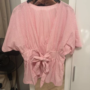 Rosa- och vitrandig topp med knyt - Superfin kort klänning med rosa och vita smala ränder. Modellen har korta ärmar, markerad midja med knytband bak och lätt volang nedtill. Perfekt för sommarens varma dagar och har en luftig, avslappnad passform.