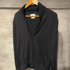 These Glory Days Merinoull fullzip - Stilren marinblå kofta från These Glory Days i mjuk och exklusiv merinoull. Koftan har hel dragkedja framtill, hög krage och ribbade muddar. Perfekt för lager-på-lager och passar till allt. Enkel och tidlös design som funkar året runt.