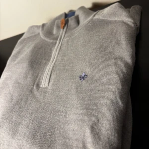 Grå stickad half zip-tröja med brodyr - Snygg grå stickad tröja från Morris med halv dragkedja och blå broderad lilja på bröstet. Tröjan har ribbade muddar och krage, perfekt för lager-på-lager. Klassisk och stilren modell som funkar året runt. Och den har tre håll men dem syns knappt så skriv bara om ni är intresserade 😁