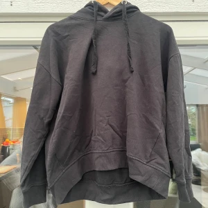 Svart hoodie med huva och snörning - En svart hoodie med klassisk huva och snörning. Tröjan har lång ärm, ribbade muddar och en avslappnad passform. Perfekt för chill dagar eller när du vill ha en enkel och stilren look.