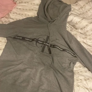 Grå Calvin Klein hoodie XL - Säljer en grå hoodie från Calvin Klein Jeans i storlek XL. Tröjan har stor CK-logga och svart band med text över bröstet, samt broderad CK-detalj på ärmen. Klassisk känguruficka och huva med snörning. Perfekt för chill dagar.