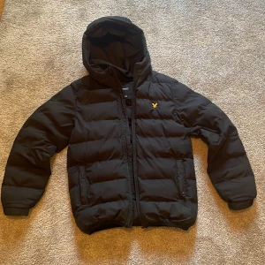 Svart pufferjacka från Lyle & Scott - Svart pufferjacka från Lyle & Scott med huva och dragkedja. Jackan har det klassiska gula logobroderiet på bröstet och två sidofickor. Perfekt för kyliga dagar och riktigt skön att bära.