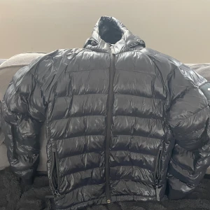 Peak Performance Svart pufferjacka med huva - Säljer en svart pufferjacka med glansig finish och huva. Jackan har dragkedja framtill och två sidofickor med dragkedja. Perfekt för kalla dagar när du vill hålla värmen och ändå se stilren ut. Hel och ren samt felfri. 