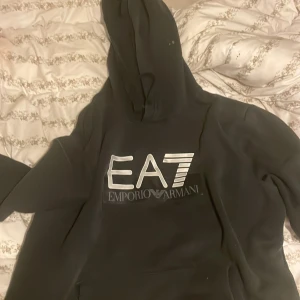 Svart EA7 Emporio Armani hoodie - Svart hoodie från EA7 Emporio Armani med stor logga framtill. Klassisk huva och känguruficka. Mjuk insida och stilren look, perfekt för dig som gillar streetwear och vill ha något snyggt och bekvämt. Lite små fläckig på 2 ställen 