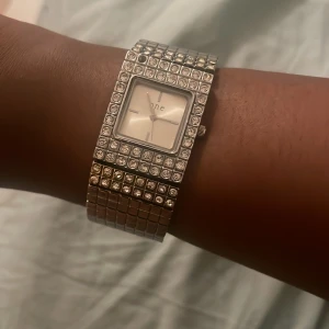 Glittrig silverklocka från O.N.E - Säljer en riktigt snygg och glittrig klocka från O.N.E med rektangulär urtavla och bred länk i silverfärgat metall. Klockan är dekorerad med massor av gnistrande stenar runt urtavlan och på armbandet. Perfekt för dig som gillar att sticka ut med accessoarer.