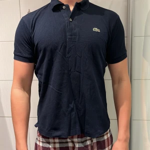 Lacoste piké  - Snygg mörkblå pikétröja från Lacoste med klassisk krage, knäppning framtill och den ikoniska krokodilloggan på bröstet. Tillverkad i bomull för en skön och luftig känsla. Använd ett fåtal gånger pga den var förliten. Skick 10/10 passar 175-180