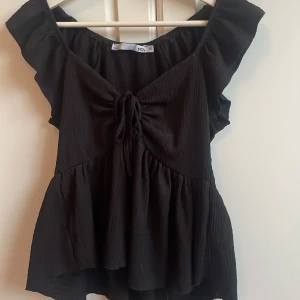 Svart blus med volang och knyt från lager 157 - Supersöt svart blus med volangärmar och knyt framtill. Blusen har en lätt rynkad struktur och är offshoulder med en luftig, flowy passform. Perfekt för dig som gillar en feminin och trendig look. Tröjan är använd några gånger och har inga synliga defekter. Kan gå ner i pris vid snabb affär 